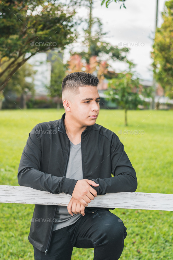 Young Latino male college student standing next to white wooden fence with hands on fence