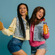 Two playful young women holding pineapple and bottle with lemonade against blue background - PhotoDune Item for Sale