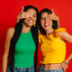 Two women in colorful wear covering eyes to each other and smiling against red background - PhotoDune Item for Sale