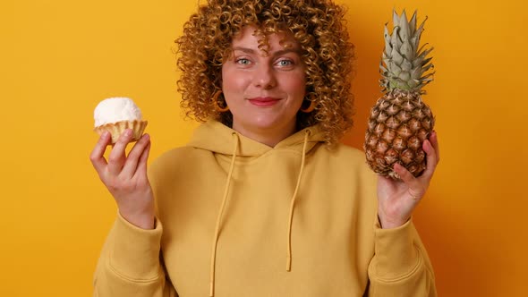 Young Woman Making Choice Between Pineapple and Tasty Sweet Cake Isolated on Yellow Color Background