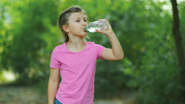A little girl drinking clean water from a bottle against the background of trees.