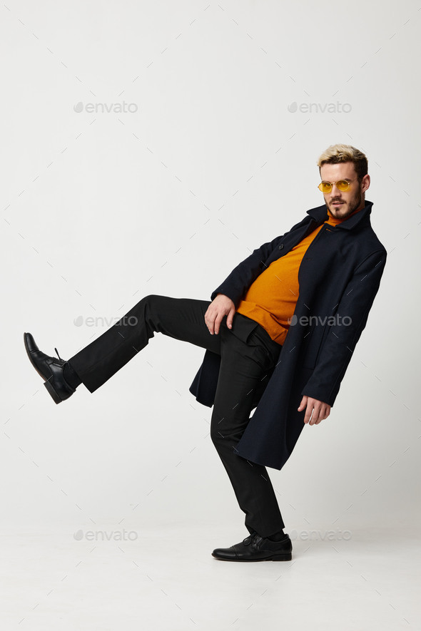 a man in an orange sweater and in a dark coat is dancing on a light background lifting his leg up