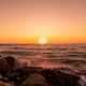 Big sun on skyline at sunset. Ocean water splash on rock beach with beautiful sunset sky and clou - PhotoDune Item for Sale