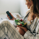 A pregnant woman sitting in pajamas eats salad and uses a mobile phone to communicate. - PhotoDune Item for Sale