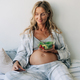 A pregnant woman sitting on the bed eats a green salad and surfs social networks on a mobile phone. - PhotoDune Item for Sale