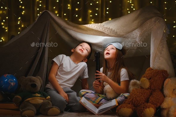 Two little child play at home in the evening to build a camping tent to read books with a flashlight