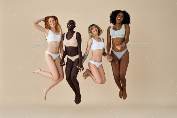 Happy diverse young women wearing underwear jumping on beige