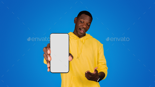 Smiling surprised young black man pointing finger at phone with blank screen, recommends app