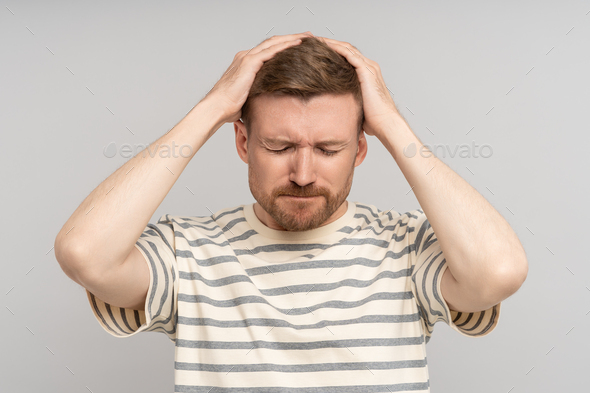 Puzzled confused bearded man holds head with closed eyes feels headache on grey background. - Stock Photo - Images