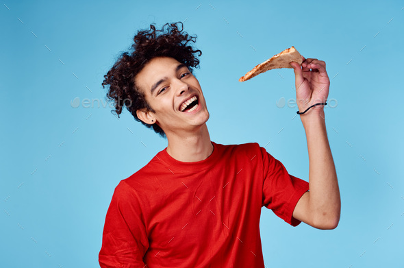 man with a piece of pizza in his hand on a blue background teenager in a red t shirt curly hair