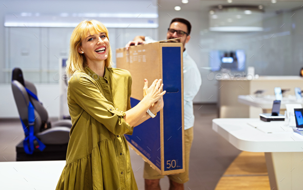 Cheerful young couple carrying packed TV they bought on sale. Tech store interior.
