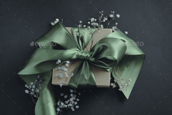 Paper gift box with olive green ribbon tied in a bow, small flowers, black  background. Stock Photo by TaniaJoy