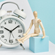 Wooden man sits near a white alarm clock. Concept of watching time, long wait. - PhotoDune Item for Sale