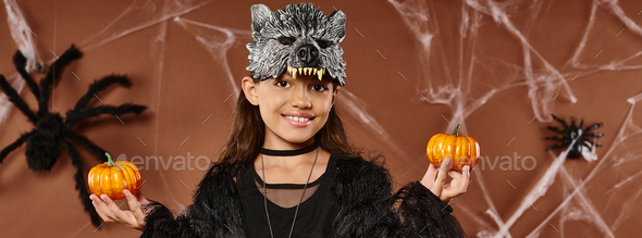preteen girl in wolf mask holds pumpkins on brown backdrop with spiders and web, Halloween, banner