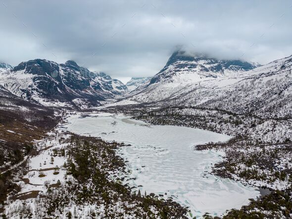 Frozen lake surrounded by snow-capped mountains in a valley on a cloudy day. Trollheimen, Norway. - Stock Photo - Images