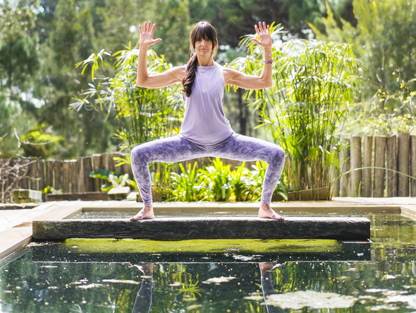 A woman practicing the yoga pose Goddess with Cactus Arms on a tree trunk over a pool.