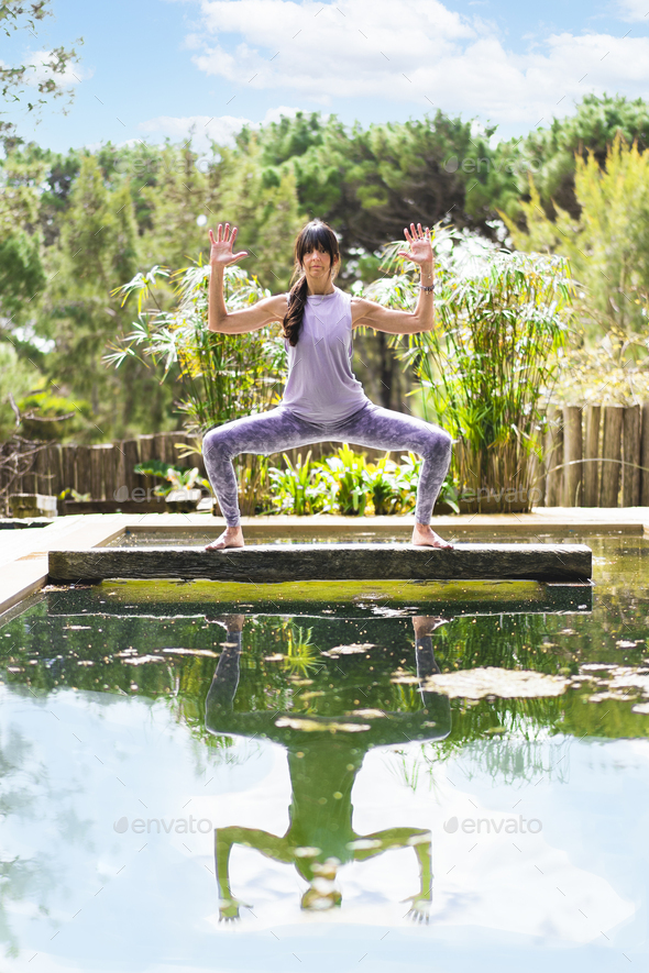A woman practicing the yoga pose Goddess with Cactus Arms on a tree trunk over a pool, Vertical shot