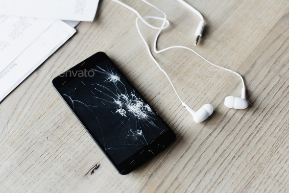 Close up view of smashed cellphone with earphones and paper sheets on wooden background