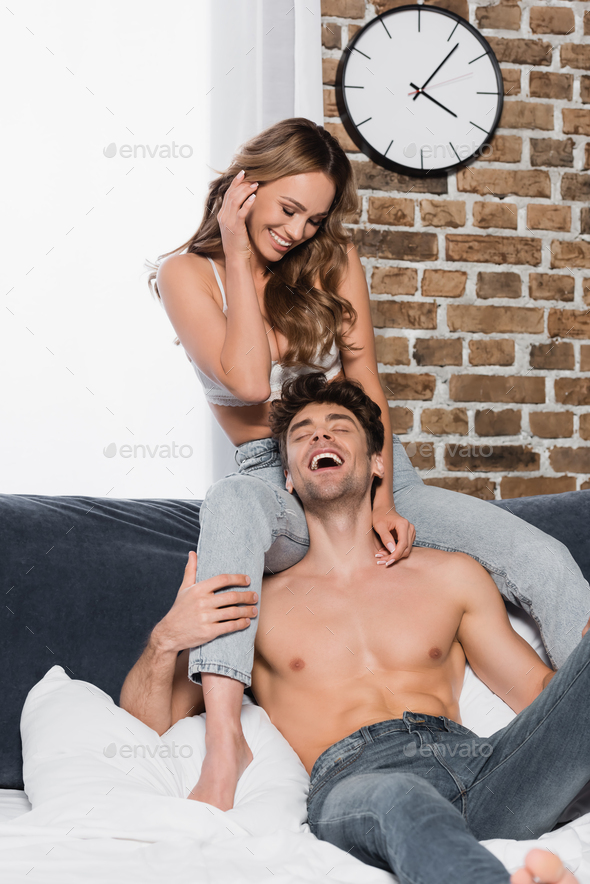attractive and smiling girlfriend in black underwear sitting on boyfriend  in bed Stock Photo by LightFieldStudios