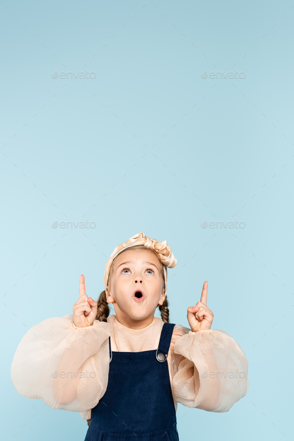 shocked kid in headband with bow pointing with fingers while looking up isolated on blue