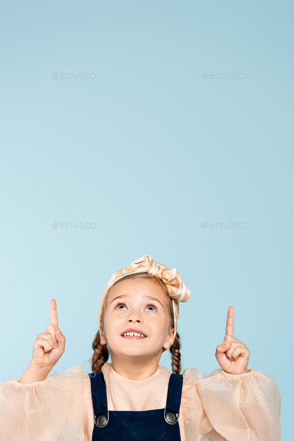 smiling kid in headband with bow pointing with fingers while looking up isolated on blue