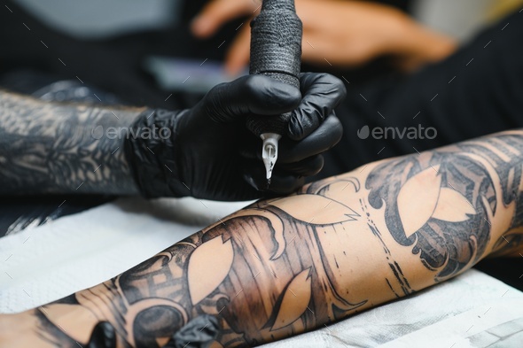 bearded tattoo artist working at his studio tattooing sleeve on the arm of his male client