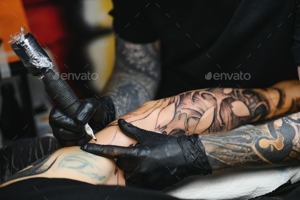 bearded tattoo artist working at his studio tattooing sleeve on the arm of his male client.