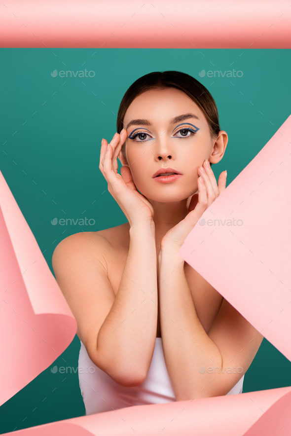 young woman with creative makeup touching face near hole in pink paper isolated on green