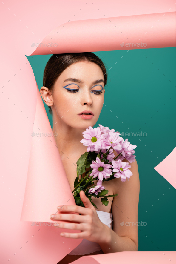 pretty woman with creative makeup holding chrysanthemums near hole in pink paper isolated on green