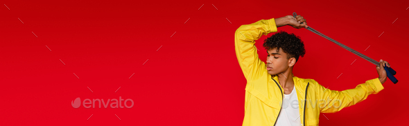 pierced african american man in yellow jacket posing with skipping rope isolated on red, banner