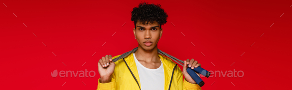 pierced african american man in yellow jacket holding skipping rope isolated on red, banner