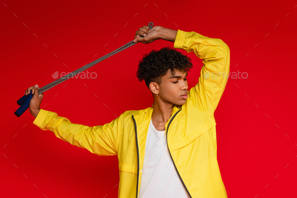 pierced african american man in yellow jacket posing with skipping rope isolated on red