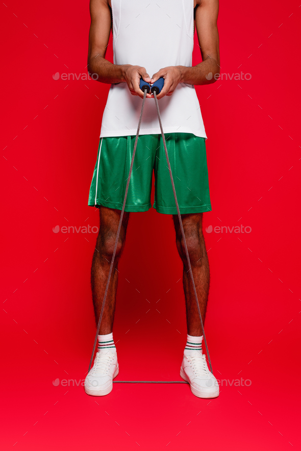 cropped view of sportive african american man in green shorts exercising with skipping rope on red