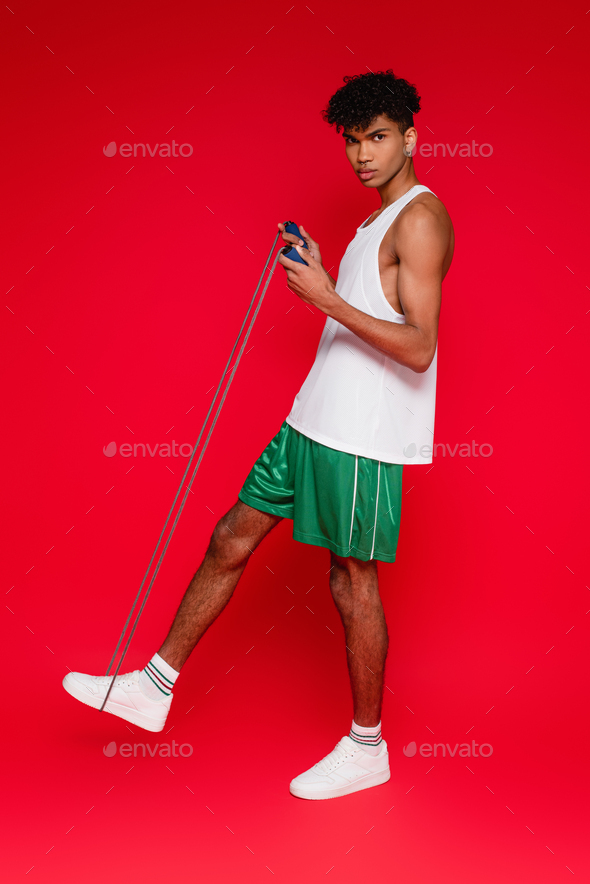 full length of sportive african american man in green shorts exercising with skipping rope on red