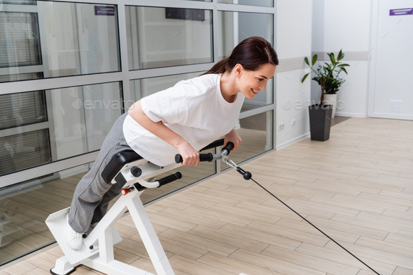 smiling woman training on pull cable exercising machine in rehabilitation center