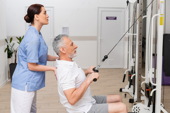 side view of rehabilitologist supporting mature man exercising on pull cable machine