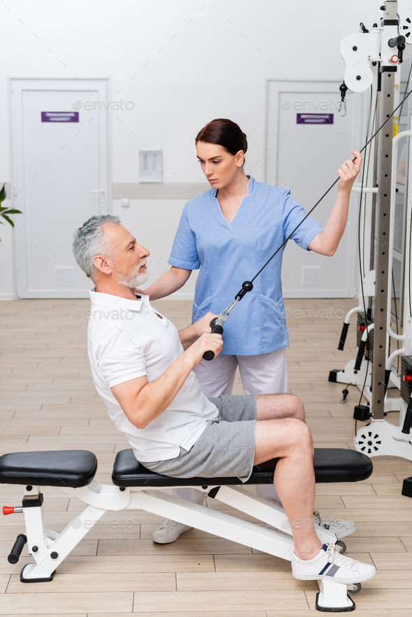 hospital trainer assisting middle aged man working out on pull cable exercising machine