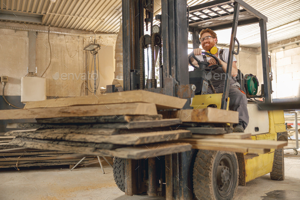Male carpenter at wooden sawmill warehouse is loading with loader wooden board