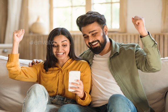 Cheerful indian couple using smartphone and shaking fists, reading great news, celebrating big luck