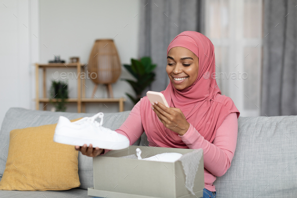 Happy black lady in hijab client unpacking parcel and taking photo of her shoes on smartphone