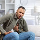 A young African-American man is suffering from a heart attack, sitting on the sofa at home and - PhotoDune Item for Sale