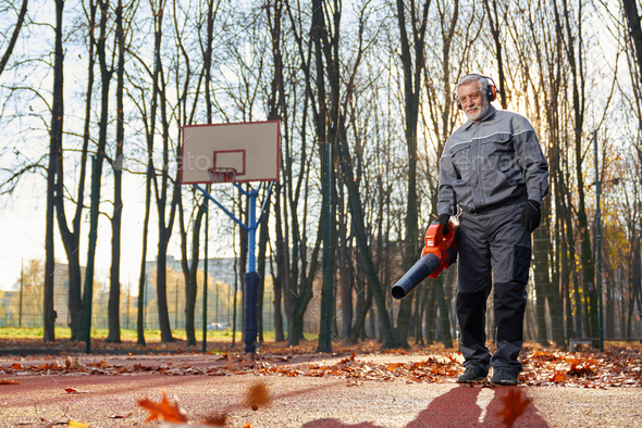 Smiling man in overalls cleaning fenced basketball court from leaves.
