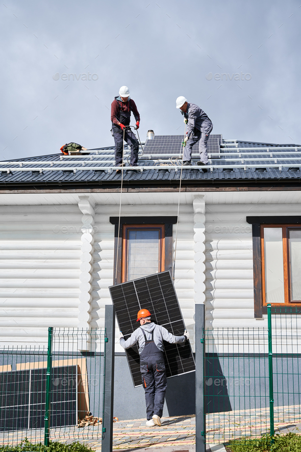 Workers lifting up photovoltaic solar module while installing solar panel system on roof of house.