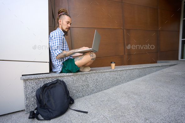 Informal guy works on laptop on granite curb near modern building