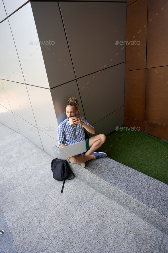 Informal guy drinks coffee on granite curb near modern building