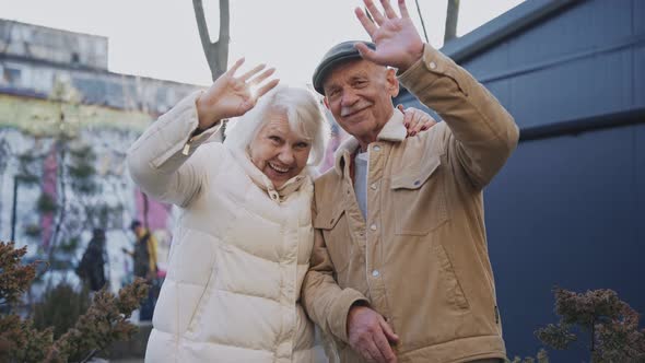 A Stylish Elderly Couple Smiling and Waving at Camera Standing on a Sunny Street