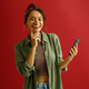 Smiling woman touching cheek while holding mobile phone over red studio background and looks camera - PhotoDune Item for Sale