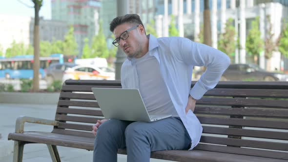 Man with Back Pain Using Laptop While Sitting Outdoor on Bench alt