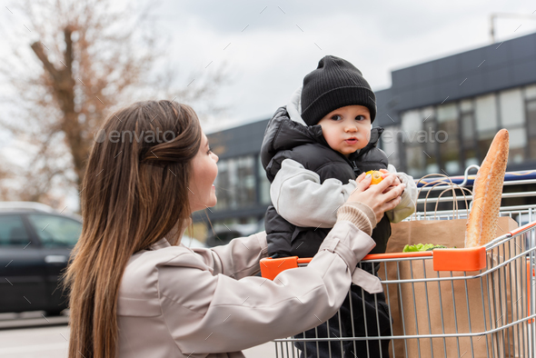 baby boy holding fresh orange while sitting in shopping cart with purchases near mother