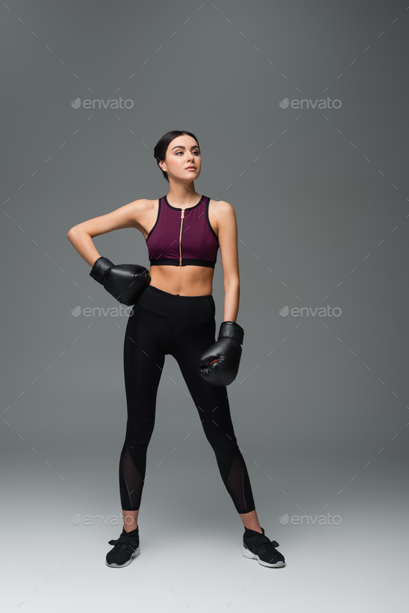 confident sportswoman in boxing gloves standing with hand on hip on grey background